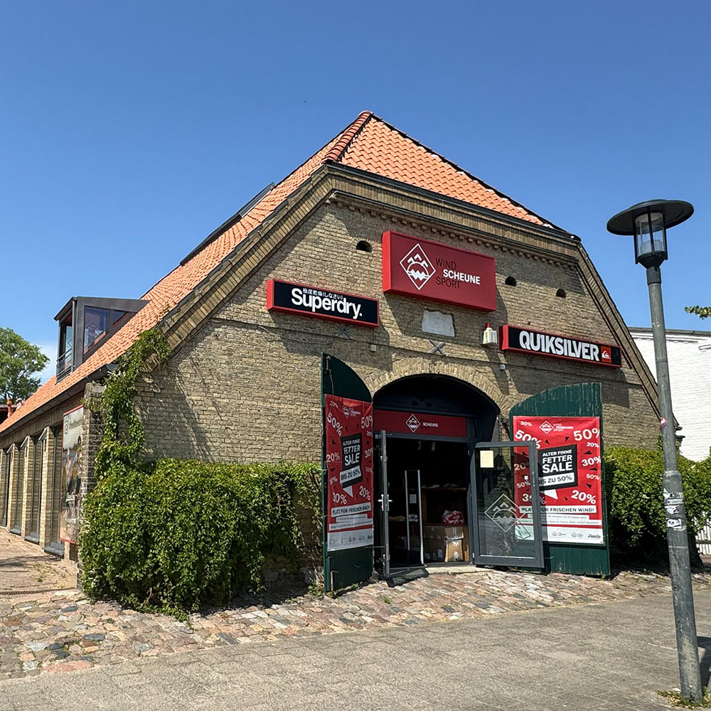 scheune1000x1000 - Windsport Fehmarn Die Backsteinfassade der Wind-Scheune mit Werbeschildern von Superdry, Quiksilver und Verkaufsplakaten im Eingangsbereich.