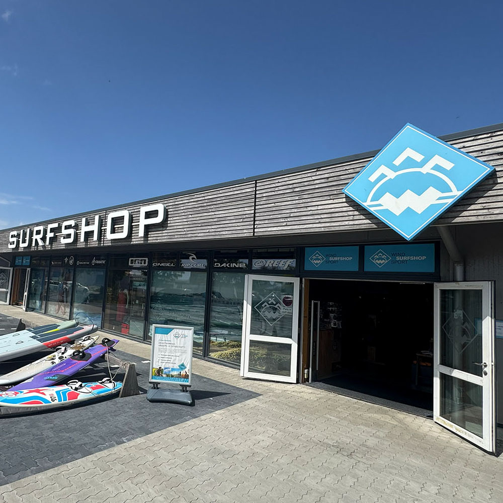 surfshop1000x1000 - Windsport Fehmarn Fassade eines Surfshops mit Surfbrettern vor dem Eingang und blauem Logo unter klarem Himmel.