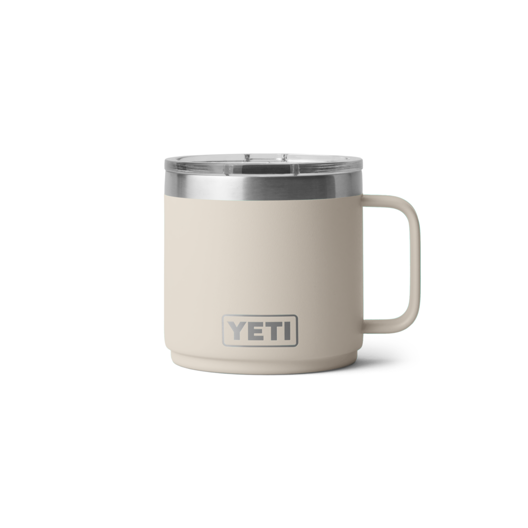 Beiger YETI Isolierbecher mit Deckel - Windsport Fehmarn Ein beiger YETI Isolierbecher aus Edelstahl mit Griff und transparentem Deckel vor weißem Hintergrund.