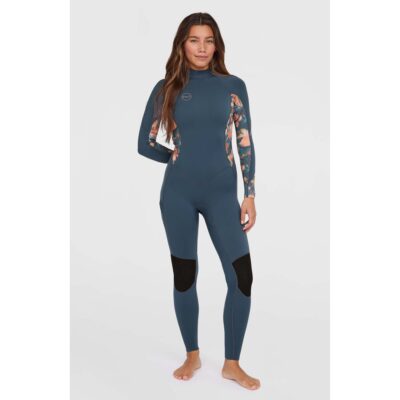 Eine Frau trägt einen blauen O'Neill Bahia Damen-Neoprenanzug mit floralem Muster an den Ärmeln vor hellem Hintergrund.