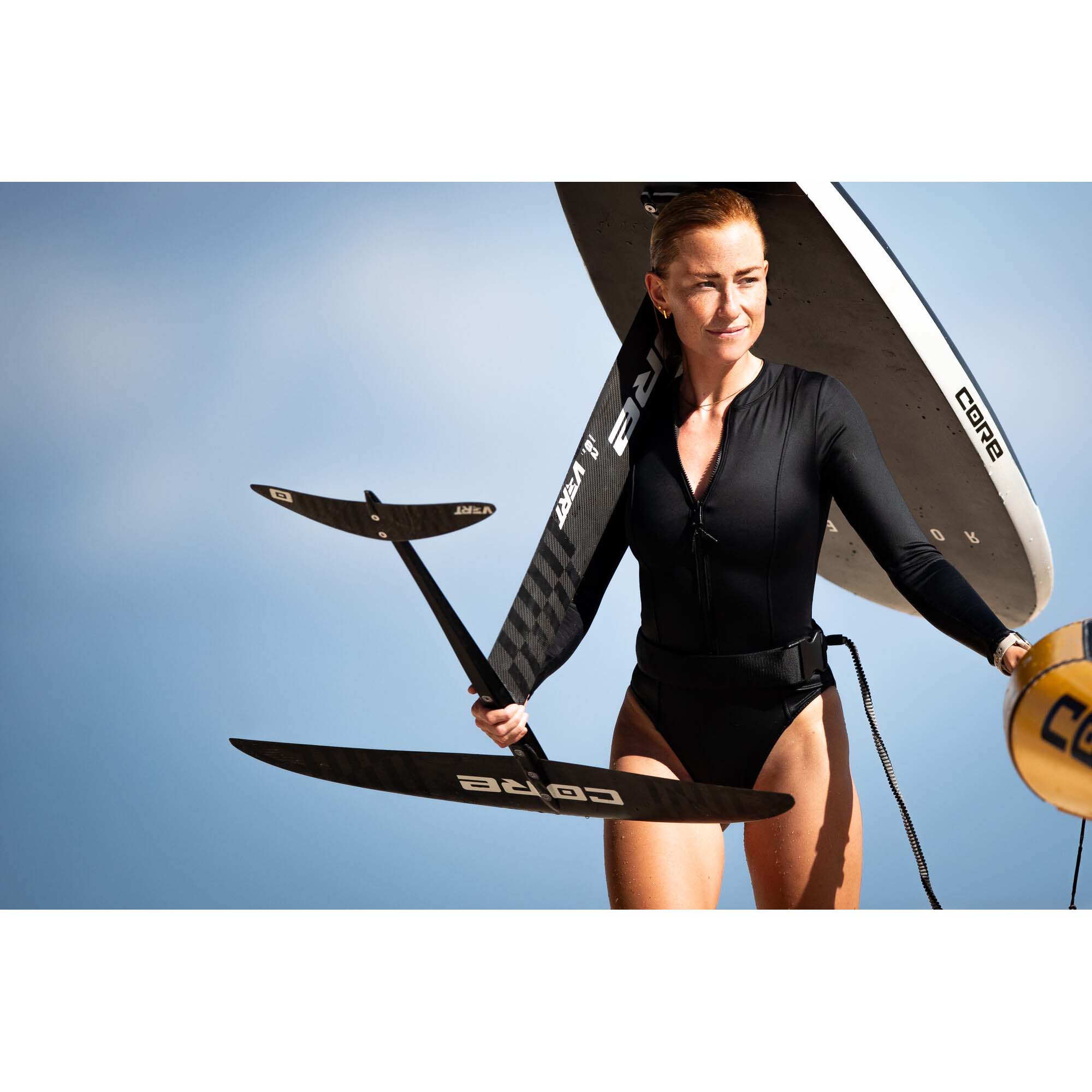 Wingfoilerin mit Core Ausrüstung - Windsport Fehmarn Eine Sportlerin trägt ein Core Wingfoil-Board und einen Foil-Mast vor blauem Himmel.