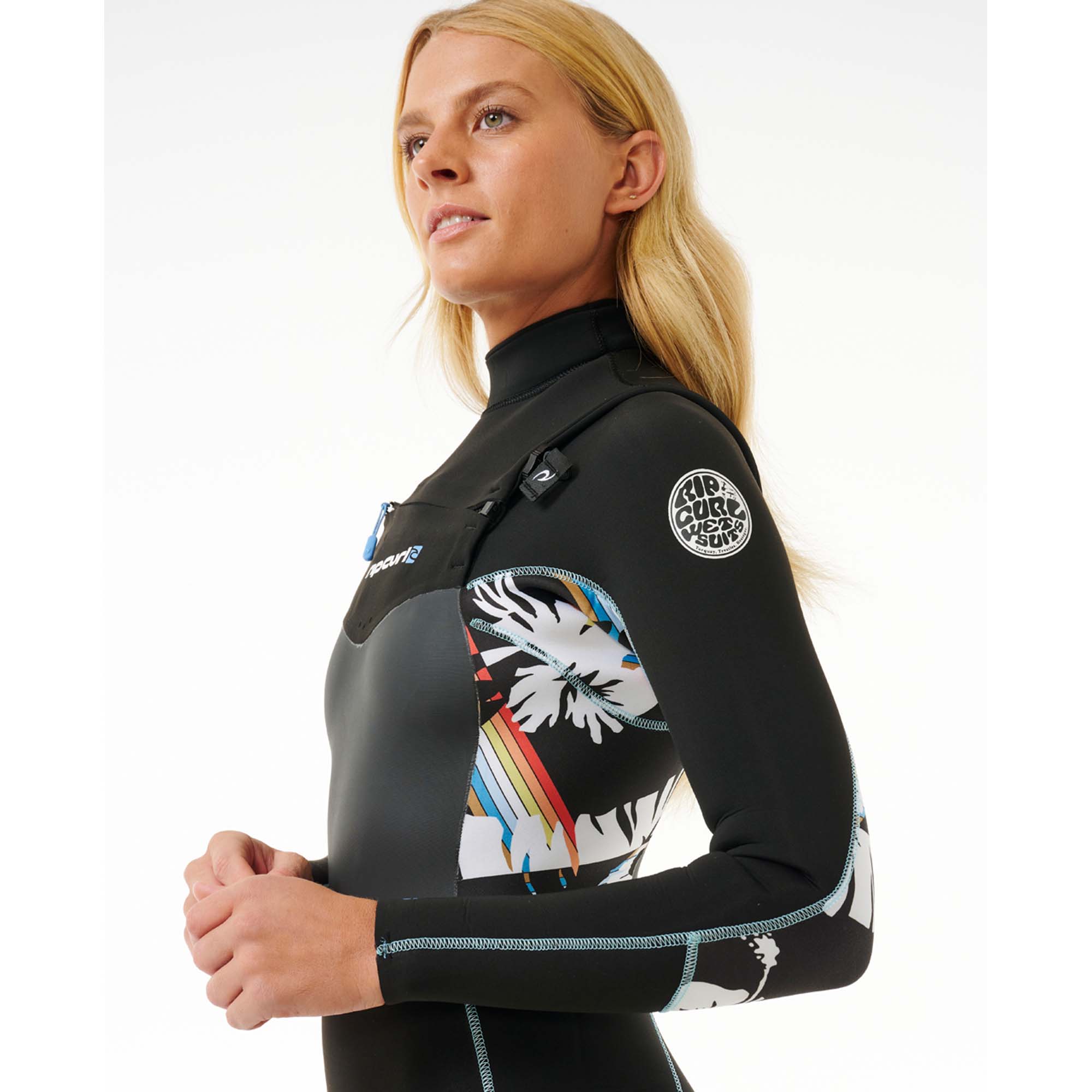 Rip Curl Surf-Neoprenanzug Damen - Windsport Fehmarn Blonde Frau im schwarzen Rip Curl Surf-Neoprenanzug mit buntem Blumenmuster an der Seite vor weißem Hintergrund.