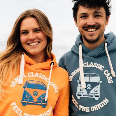Zwei Personen tragen orangene und blaue Unisex-Hoodies mit VW-Bus-Motiv und dem Schriftzug Feel The Origin.